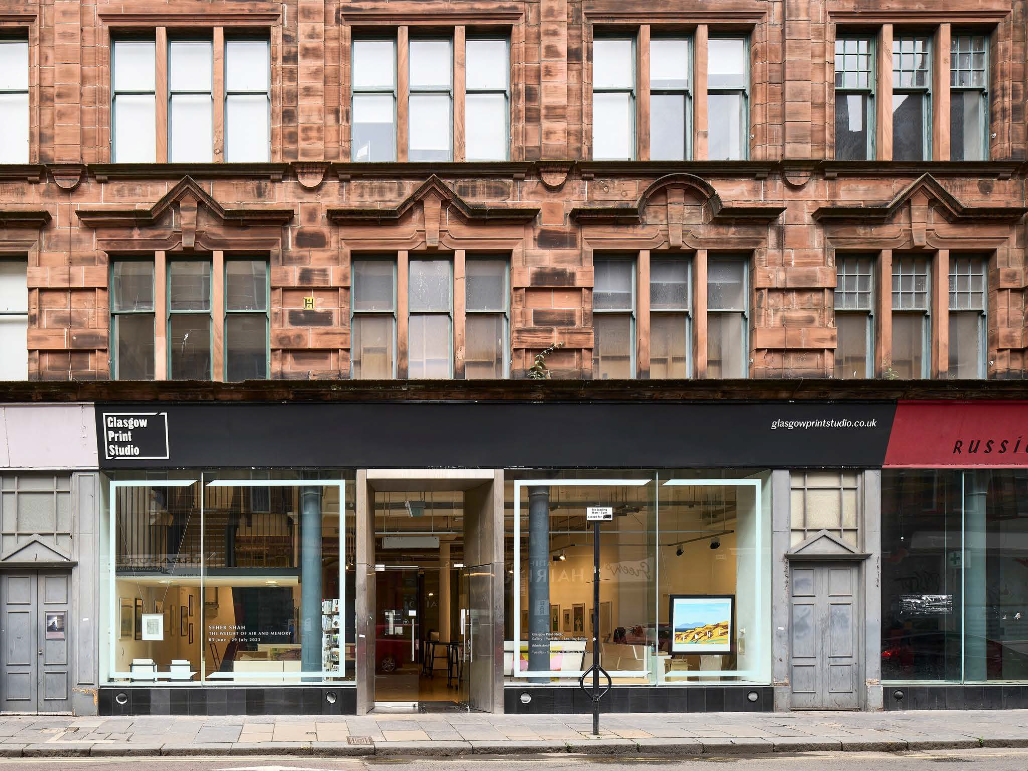 The image shows the exterior of the Glasgow Print Studio, housed in a red sandstone building with large windows and modern black signage. The studio's display windows reveal framed artworks and a glimpse into the bright gallery space inside.